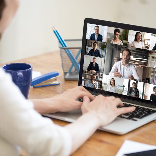Laptop screen view diverse businesspeople involved at group videocall On laptop screen web cam view diverse age and ethnicity businesspeople taking part at group videocall, view over woman shoulder sit at desk working from home. Distant communication, modern app concept